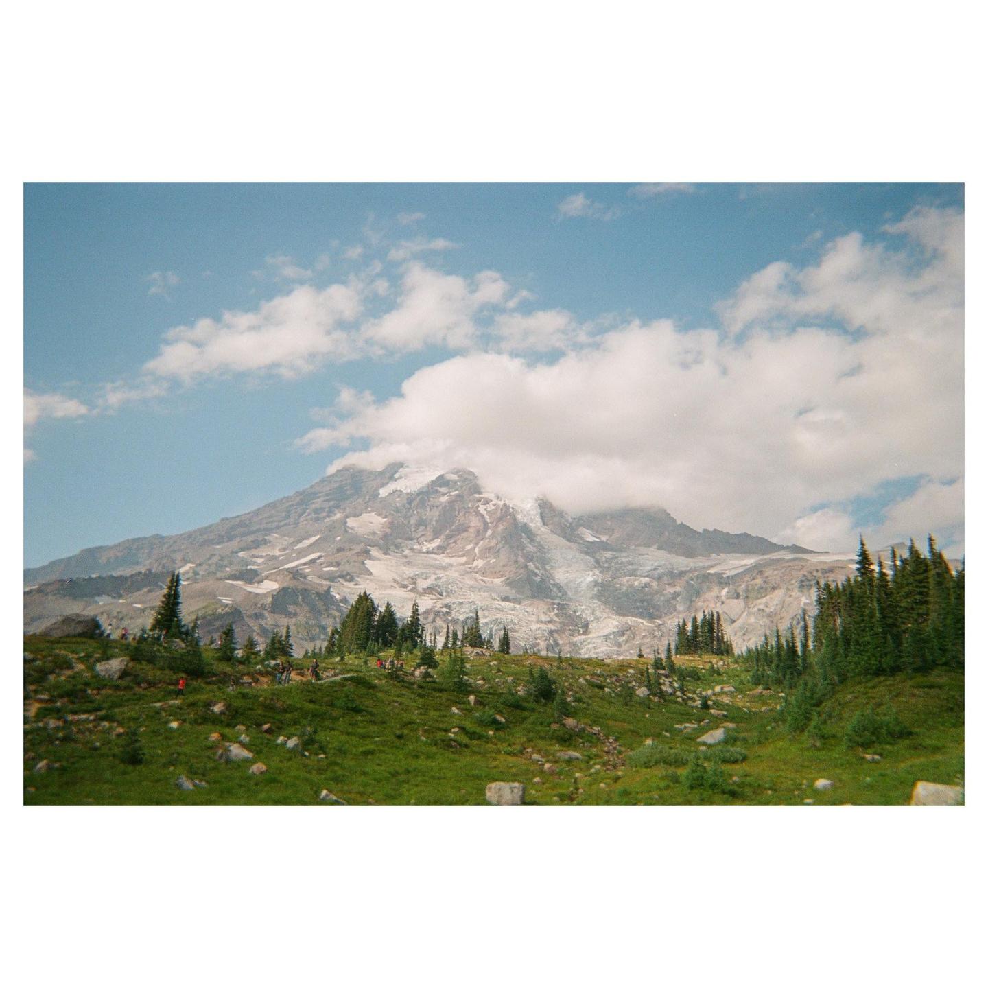 A large mountain with snow and clouds is visible behind a green, rocky hill with evergreen trees.
