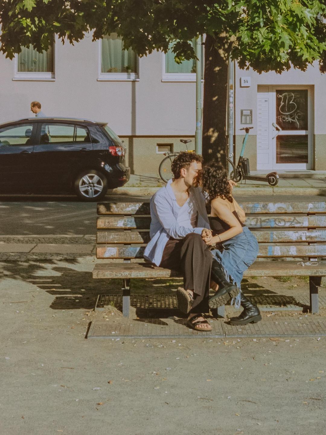 A couple sits on a park bench and kisses under a tree on a sunny day.