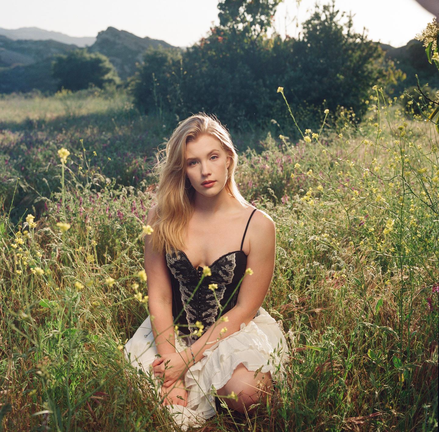 A woman kneels in a field of tall grass and wildflowers with mountains in the background.