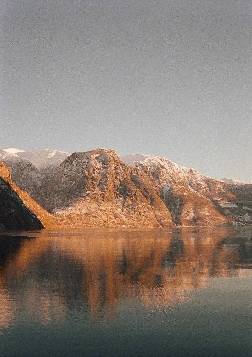 Snow-capped mountains are reflected in the still water of a fjord during golden hour.