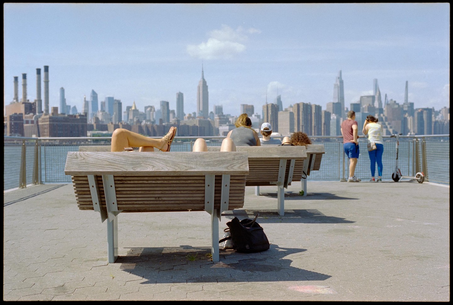 People sit on benches and stand by a railing overlooking a city skyline with a river in the foreground.