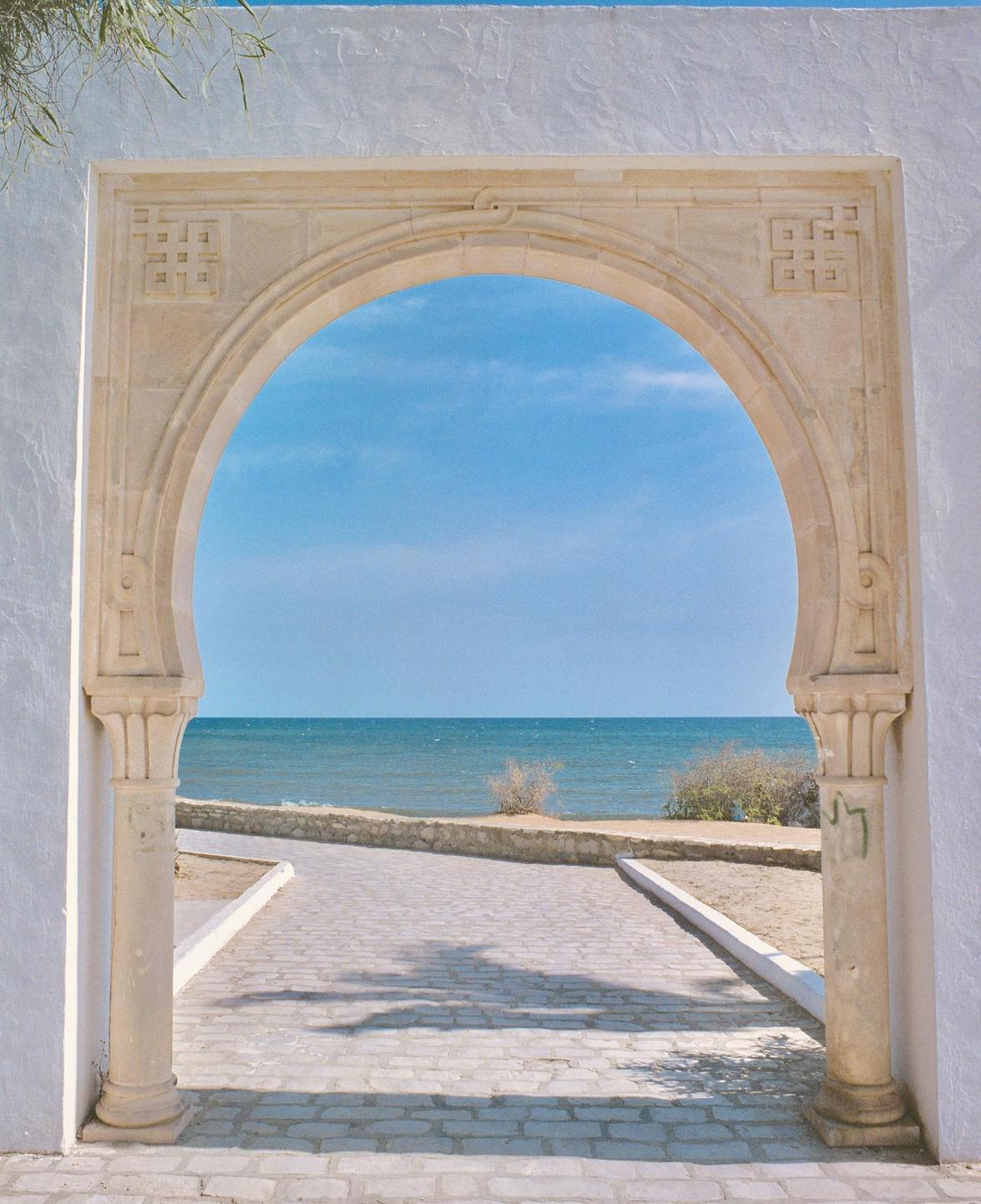 A stone archway frames a view of the ocean and a paved path leading towards the water.