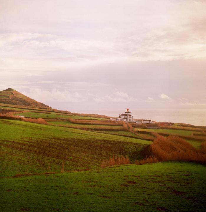 A white lighthouse with a red top stands on a green hillside overlooking the ocean under a cloudy sky.