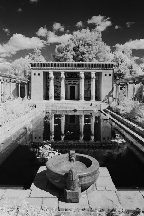 A classical building with columns is reflected in a pool of water, with a stone fountain in the foreground.