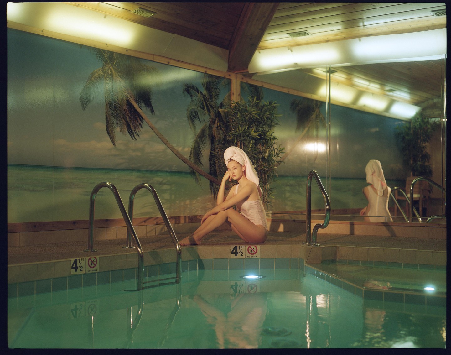 A woman wearing a towel on her head and a swimsuit sits by a swimming pool with a tropical mural in the background.