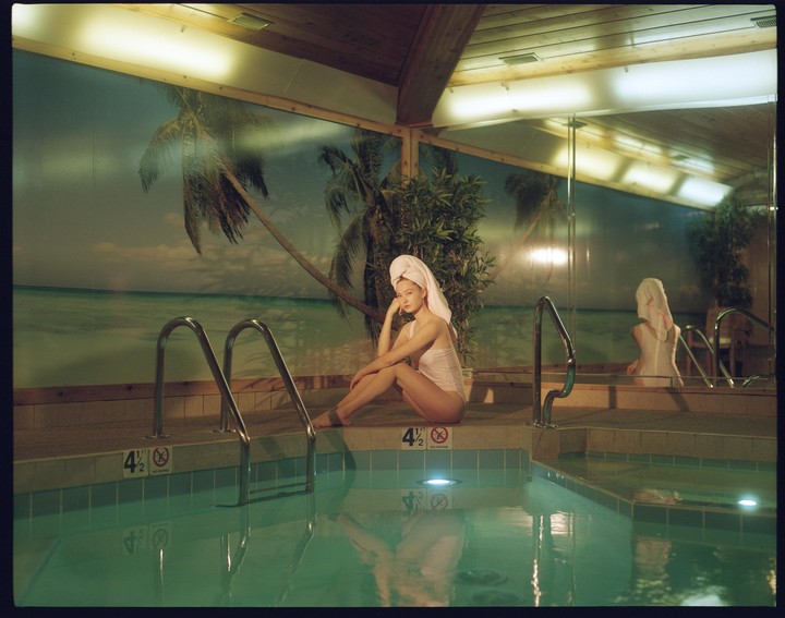 A woman wearing a towel on her head and a swimsuit sits by a swimming pool with a tropical mural in the background.