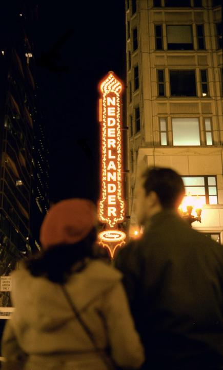 A couple stands with their backs to the camera, looking at a tall, illuminated "Nederlander" sign at night.