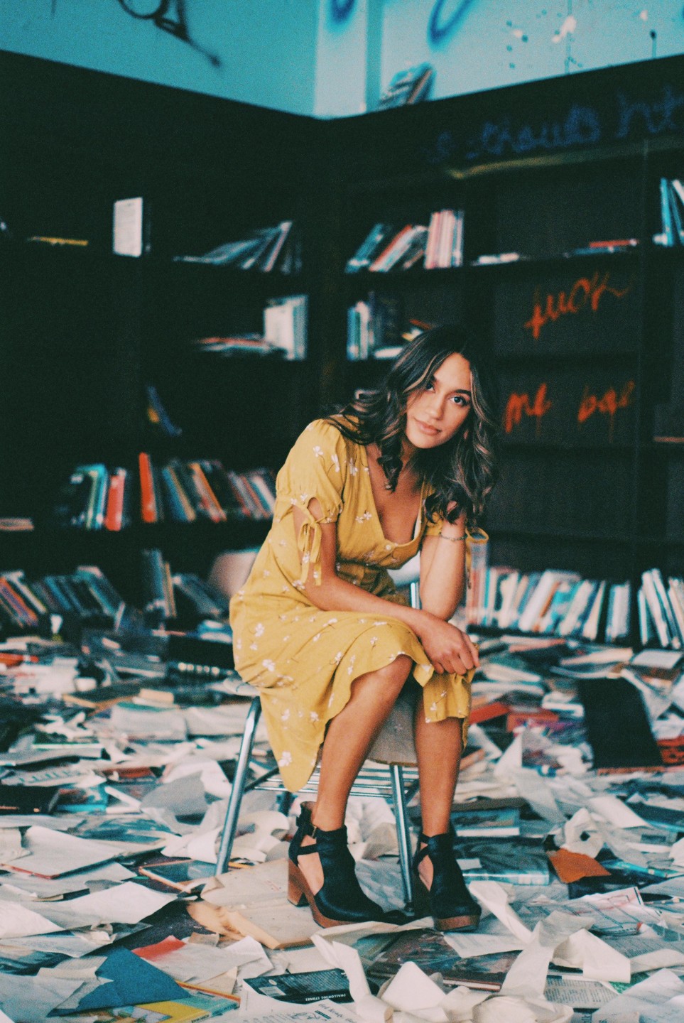 A woman in a yellow dress sits on a chair in a room with books on shelves and papers scattered on the floor.