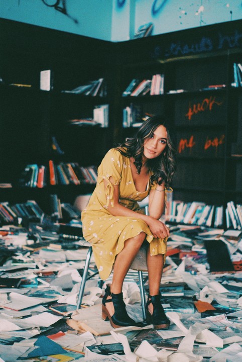 A woman in a yellow dress sits on a chair in a room with books on shelves and papers scattered on the floor.