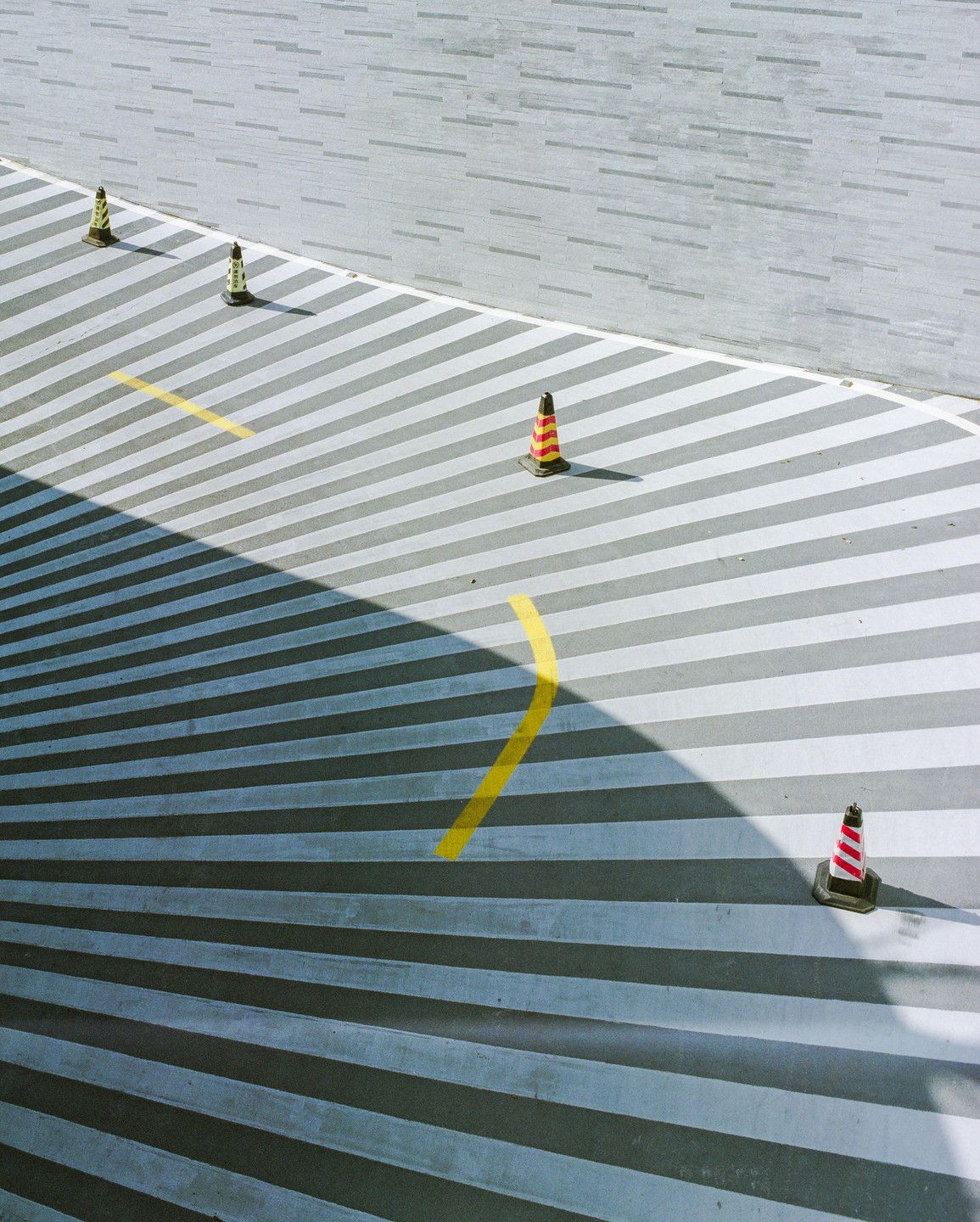 Four traffic cones are on a striped surface next to a textured wall, with a shadow cutting across the stripes.