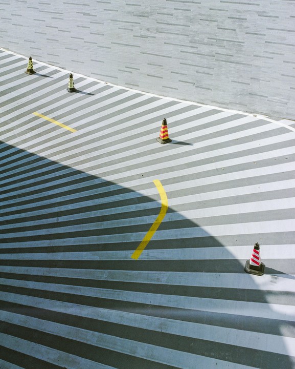 Four traffic cones are on a striped surface next to a textured wall, with a shadow cutting across the stripes.