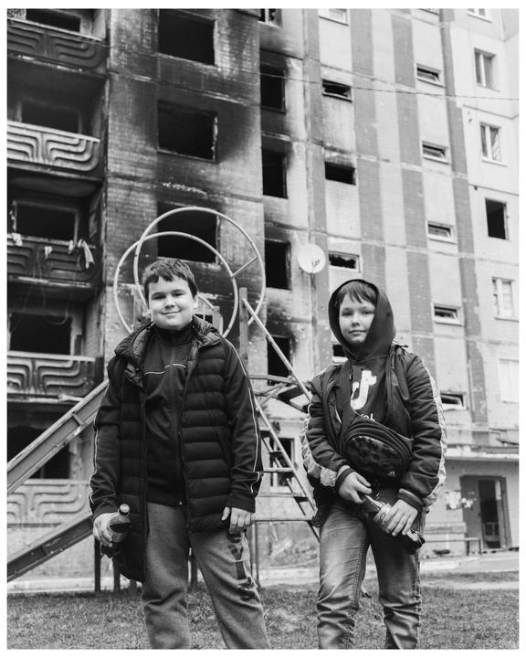 Two boys stand in front of a war-damaged building with broken windows and charred walls.