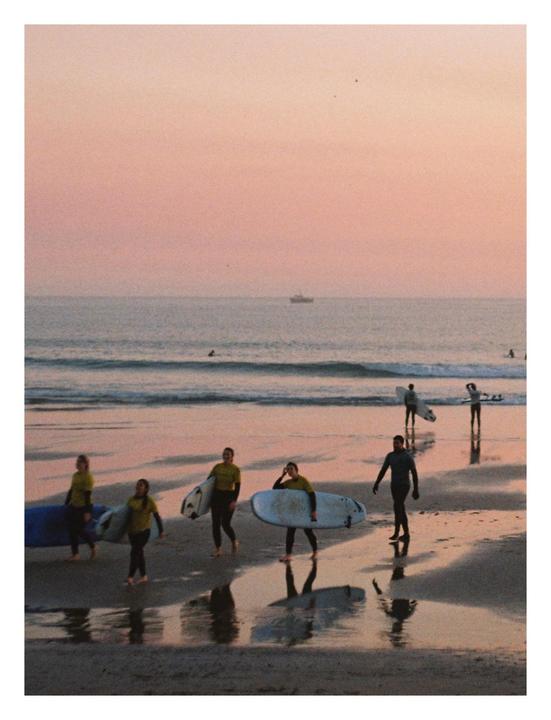 A group of surfers carrying their boards walk along a beach at sunset.