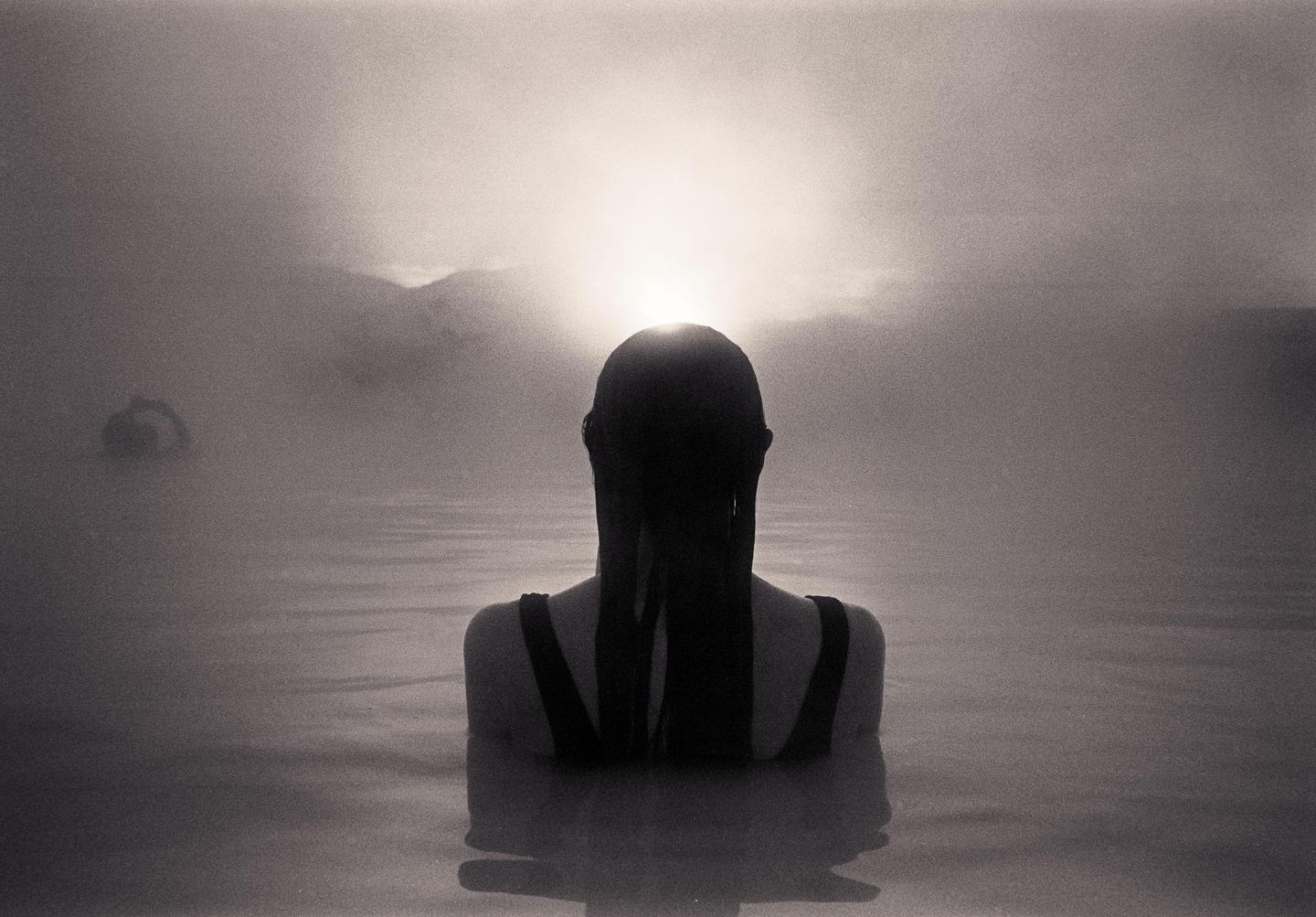 A woman with long hair is seen from behind, submerged in water with a bright light source on the horizon.