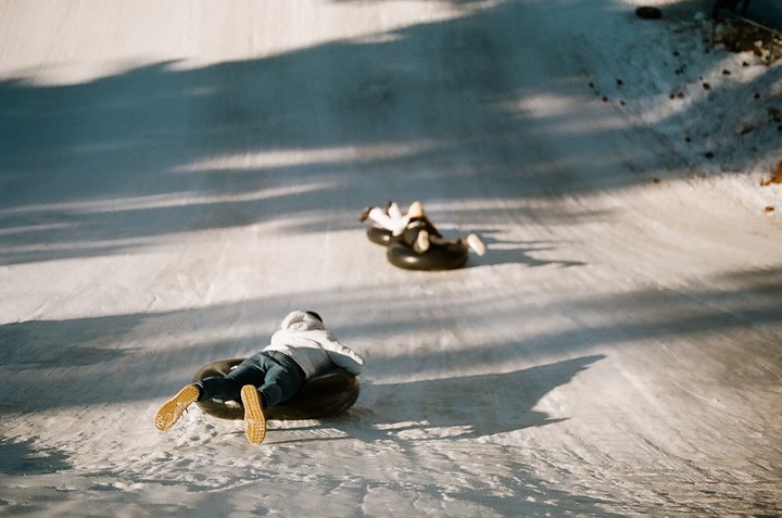 Two people slide down a snowy hill on inner tubes, casting long shadows.