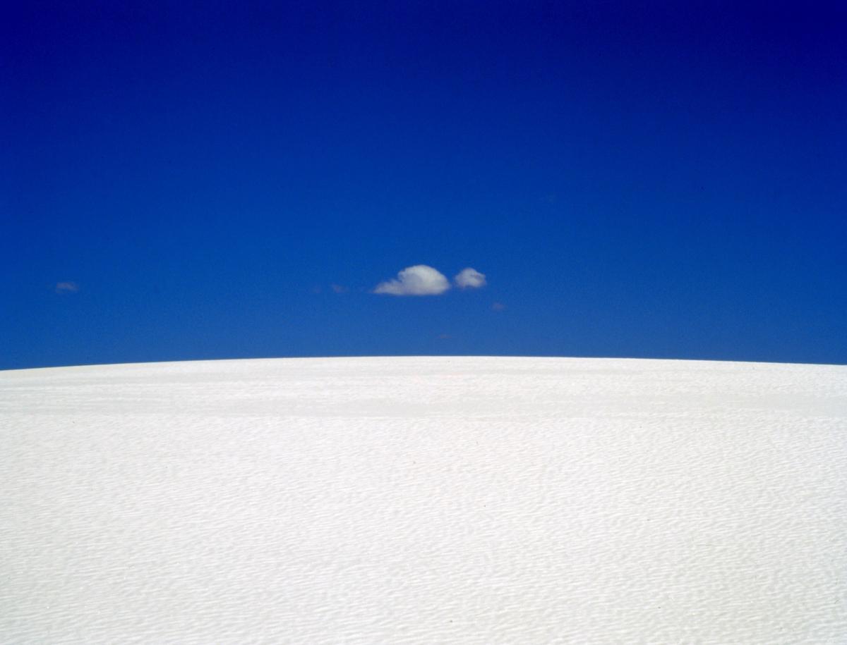 A small cloud floats in a deep blue sky above a white sand dune.