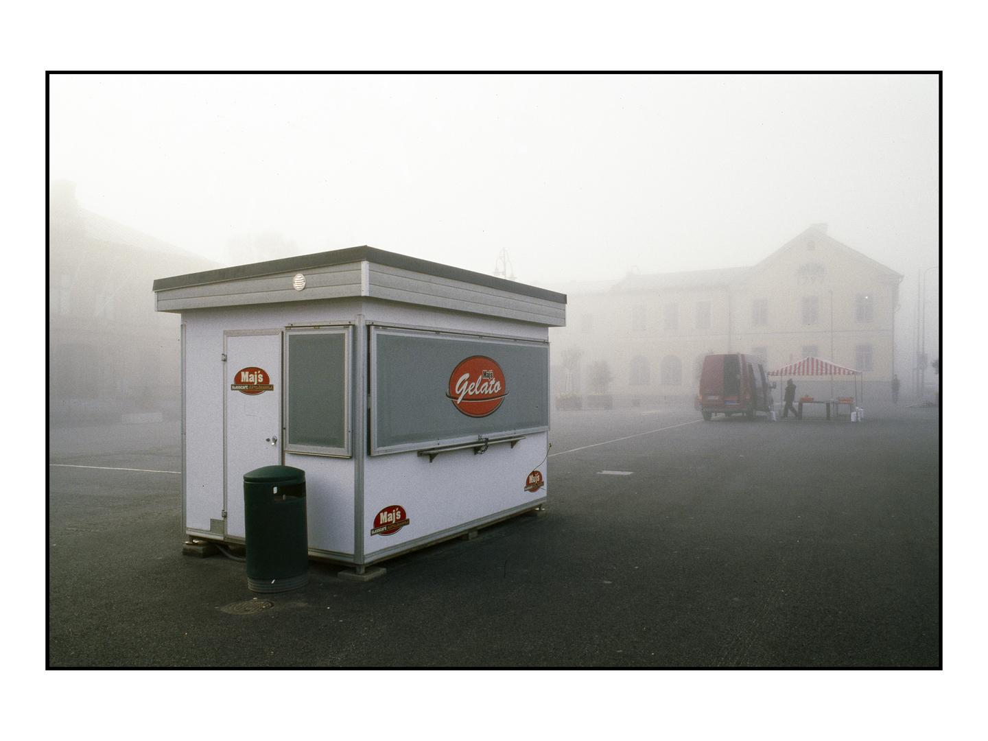 A white gelato stand with a green trash can sits in a foggy square with a red van and a striped tent in the background.