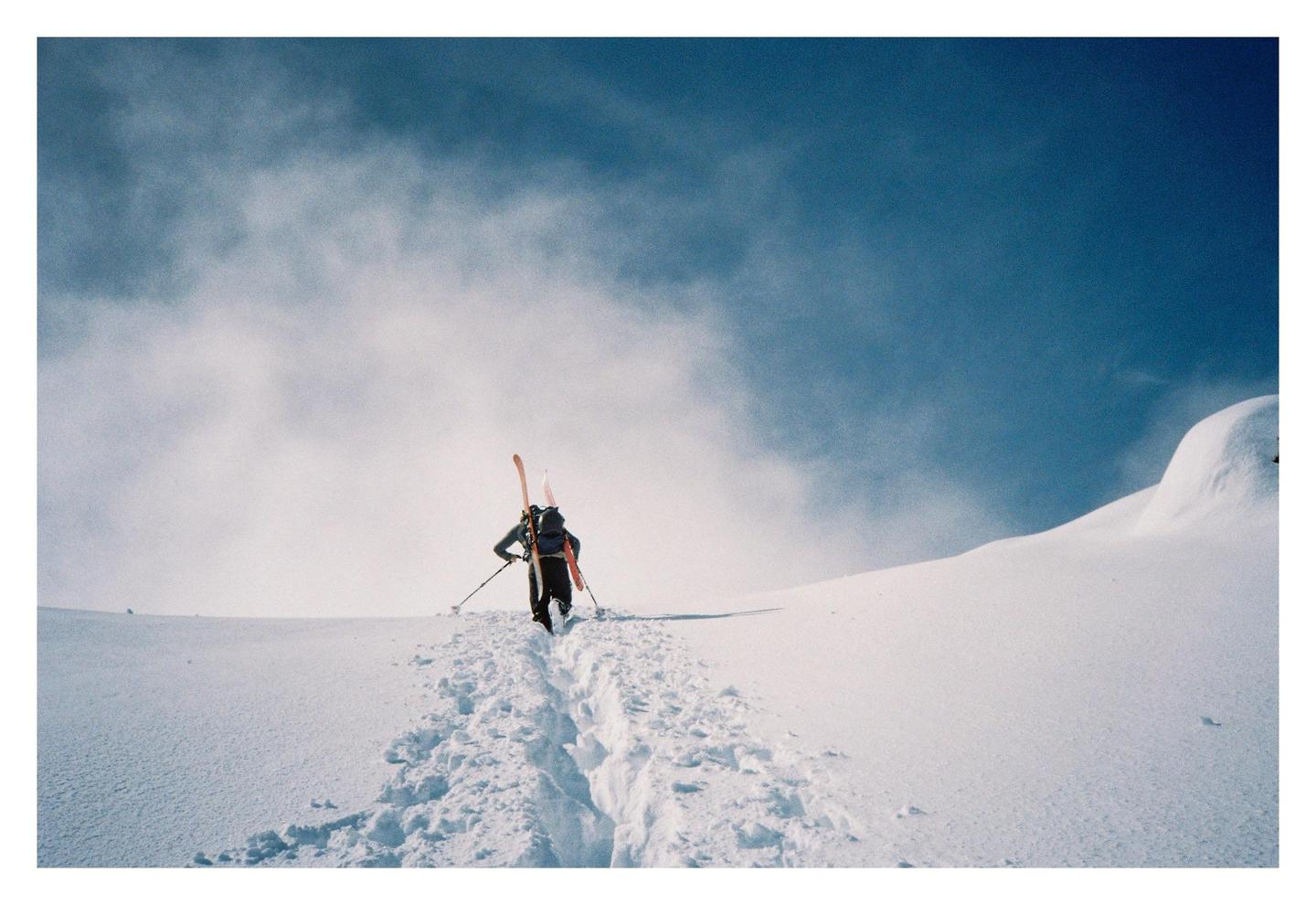 A person with skis and poles walks up a snowy mountain, leaving footprints in the snow.