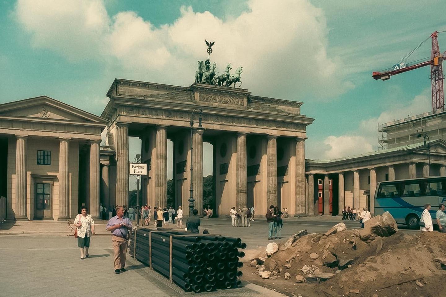 The Brandenburg Gate stands tall with a construction crane visible in the background and people walking in the foreground.