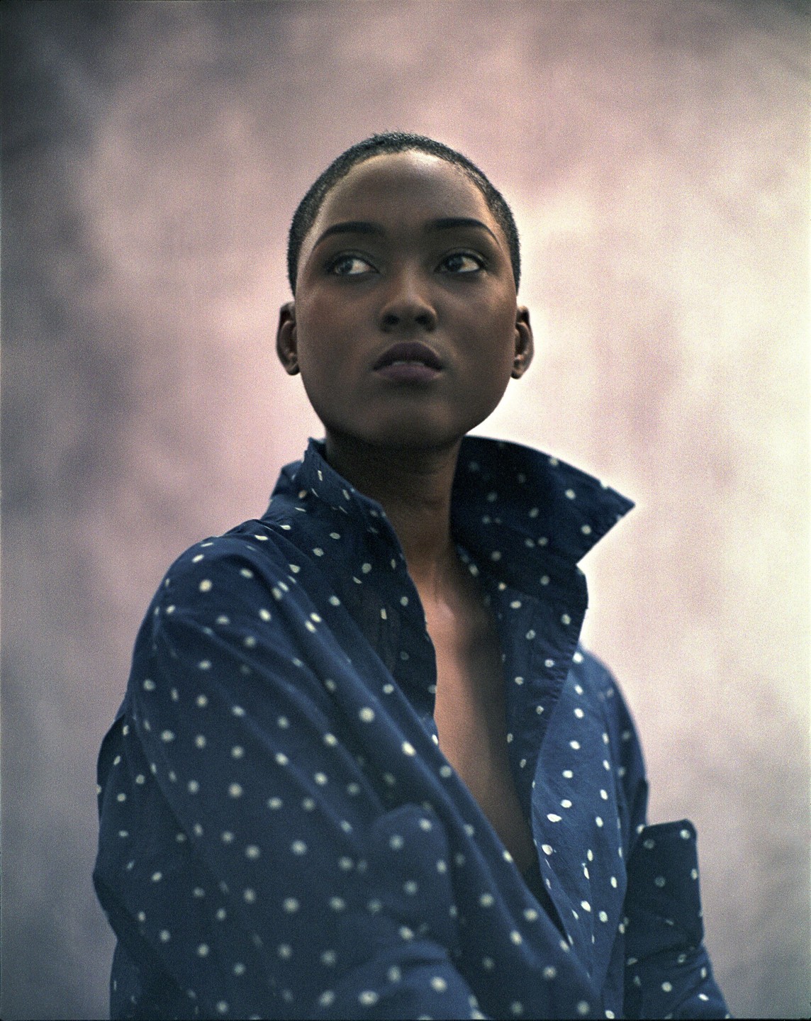 A woman with a shaved head wears a blue polka dot shirt with an open collar.