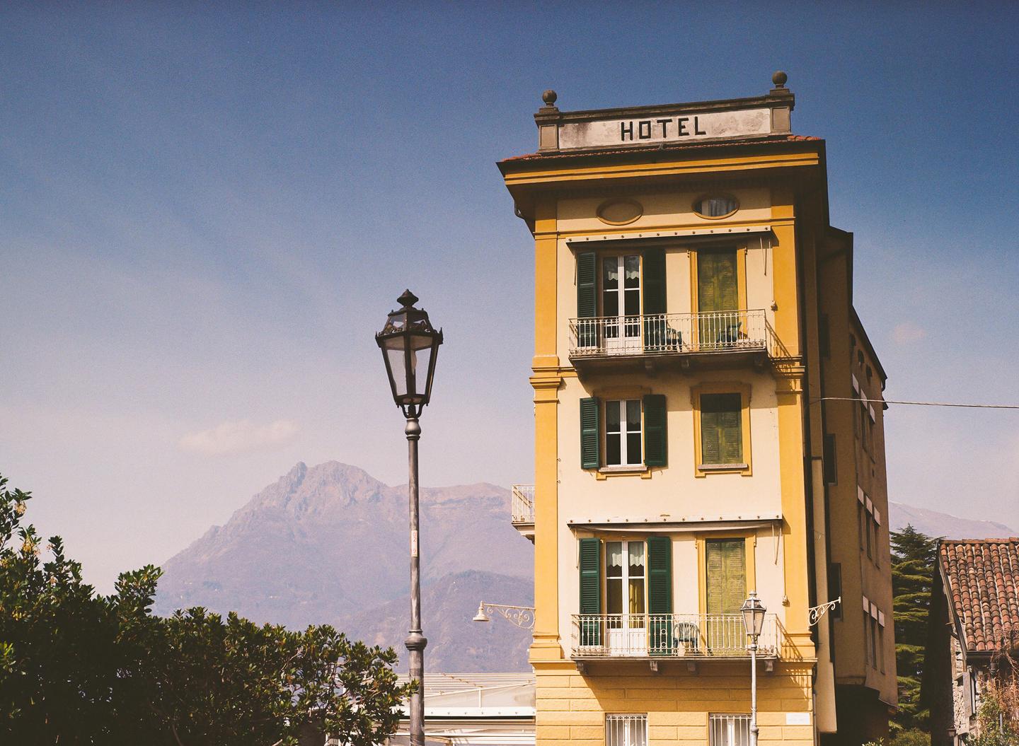 A yellow hotel building with green shutters and balconies stands next to a street lamp, with mountains in the background.