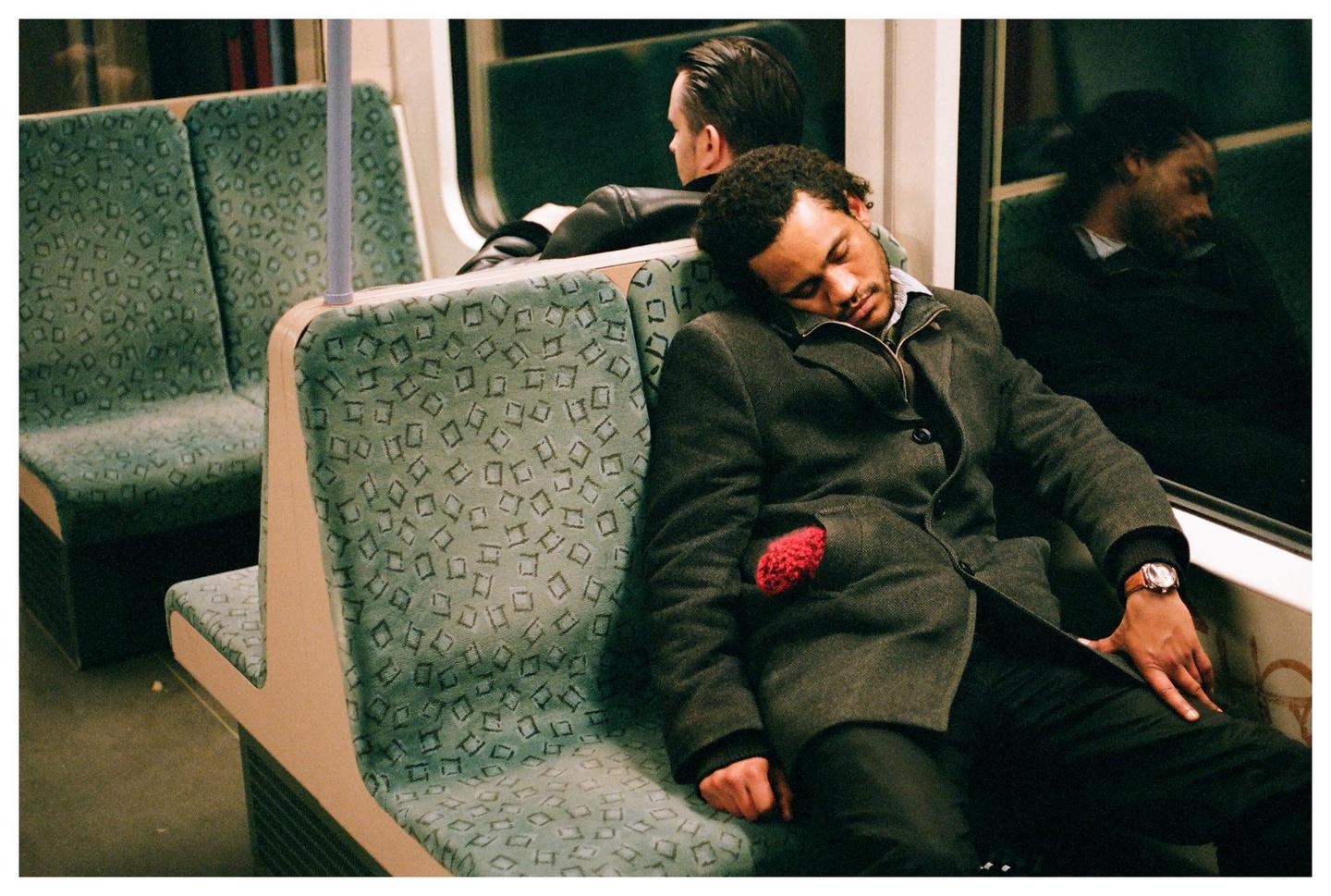 A man sleeps on a train seat next to a window, with another man visible in the background.