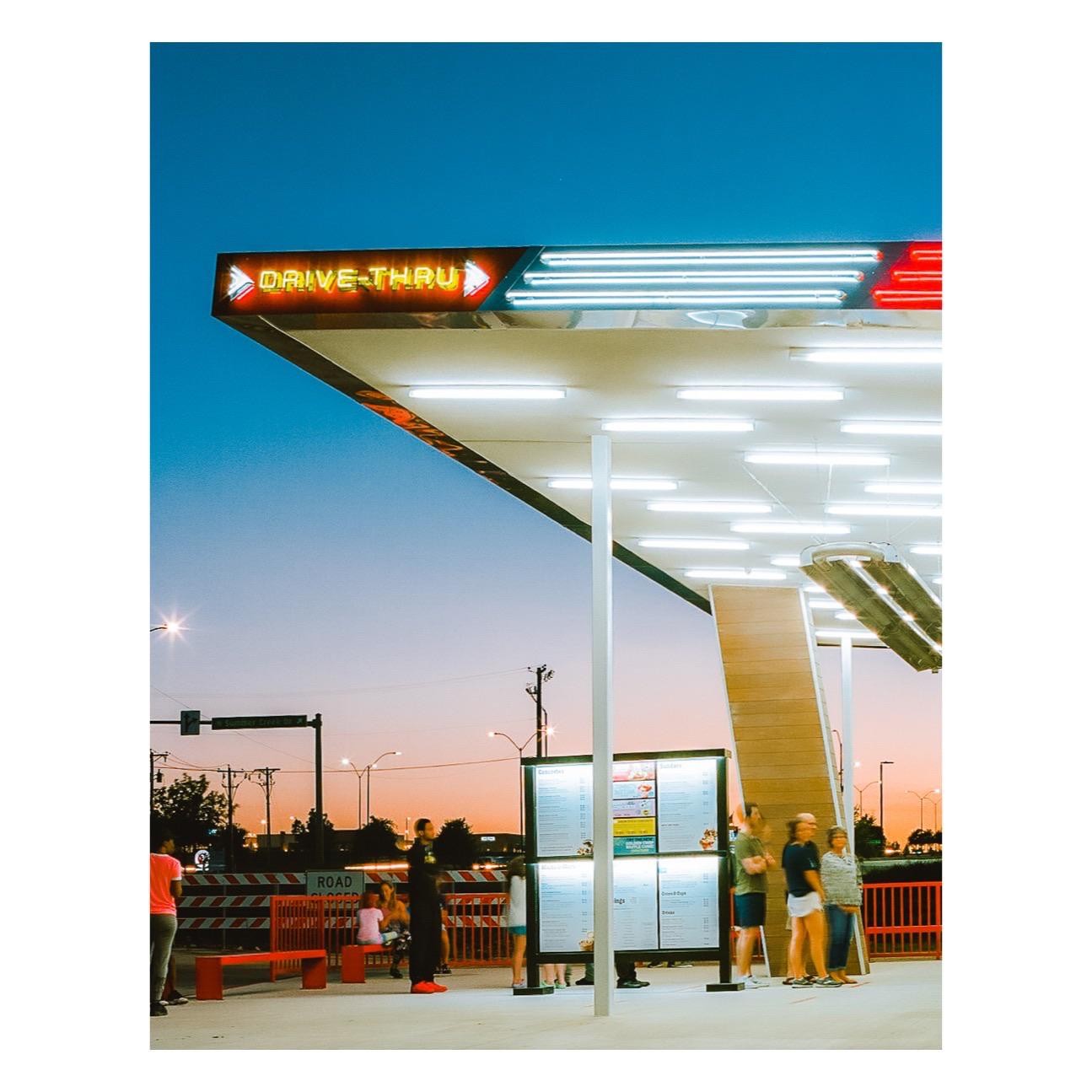 A drive-thru restaurant with a bright neon sign is illuminated at dusk.