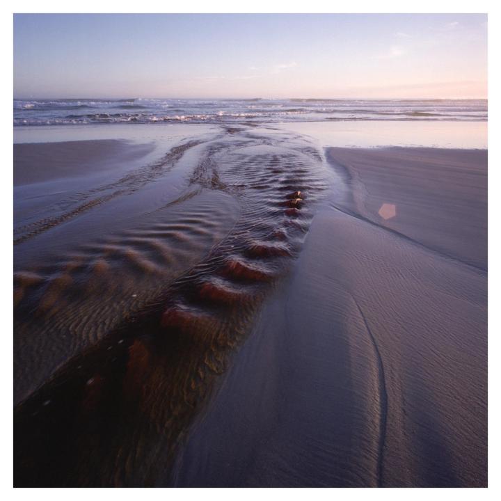 A stream of water flows across a sandy beach towards the ocean under a bright sky.