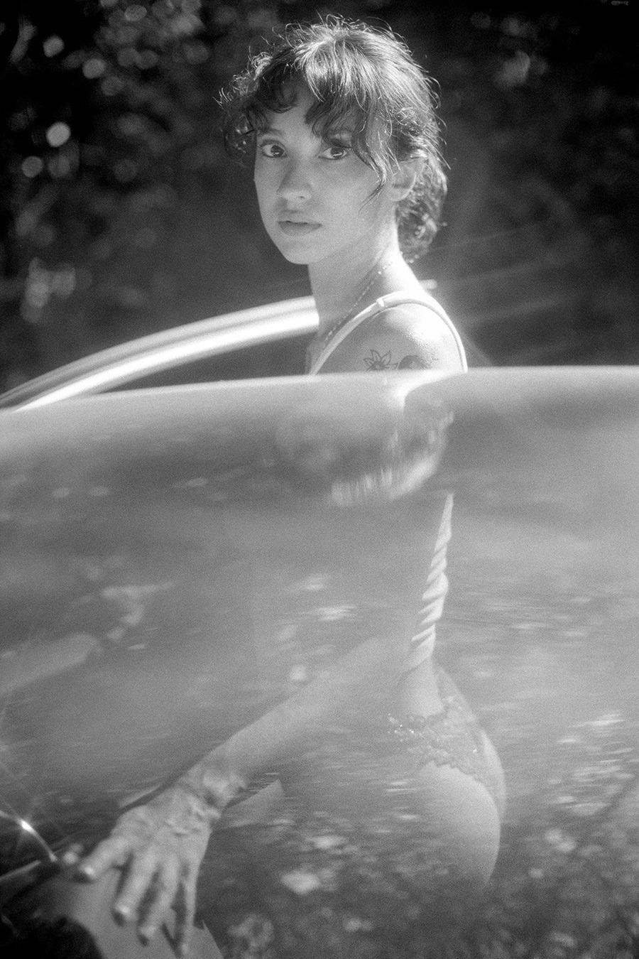 A woman with curly hair looks over her shoulder while partially submerged in water, with a car roof visible in the foreground.
