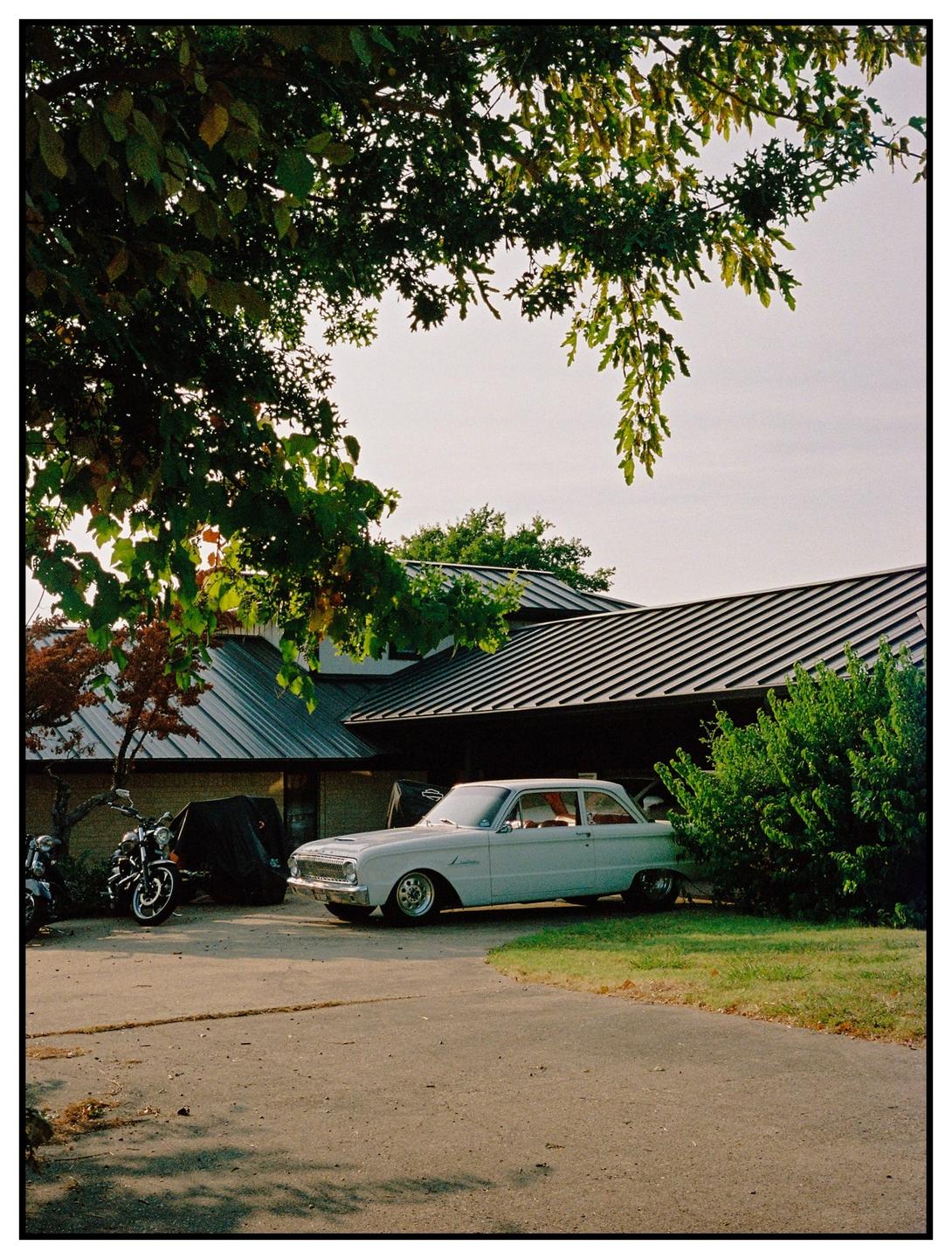 A light blue vintage car is parked in a driveway next to a house with a dark roof and green bushes.