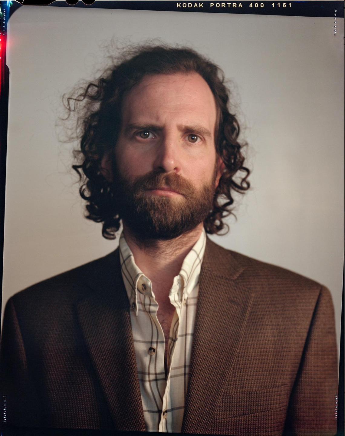 A man with curly hair and a beard wears a brown jacket and a plaid shirt.