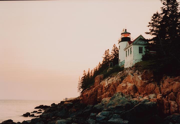 A white lighthouse with a red roof stands on a rocky cliff overlooking the ocean at dusk.