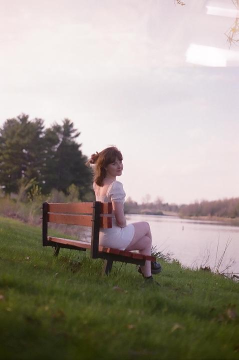 A woman sits on a park bench next to a body of water, looking over her shoulder at the viewer.