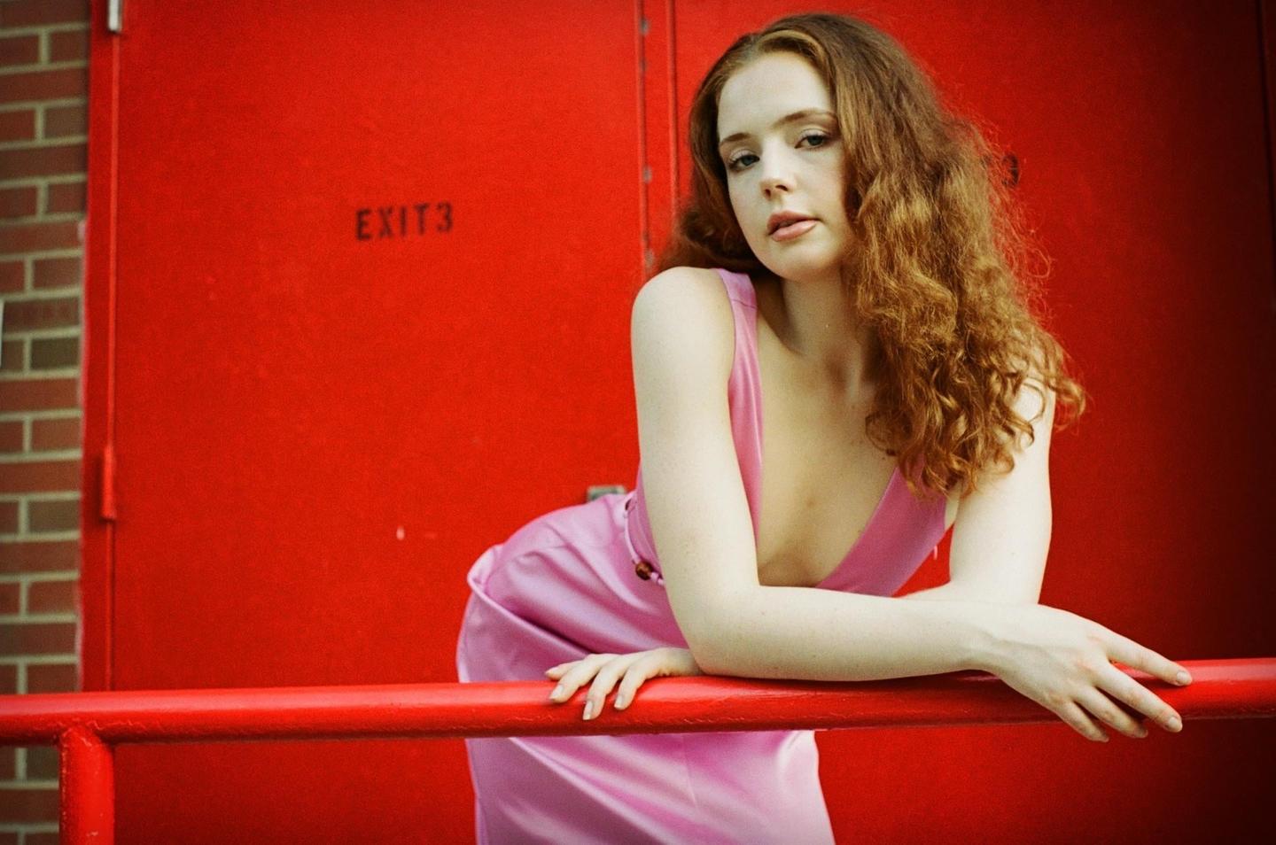 A woman in a pink dress leans on a red railing in front of a red door.