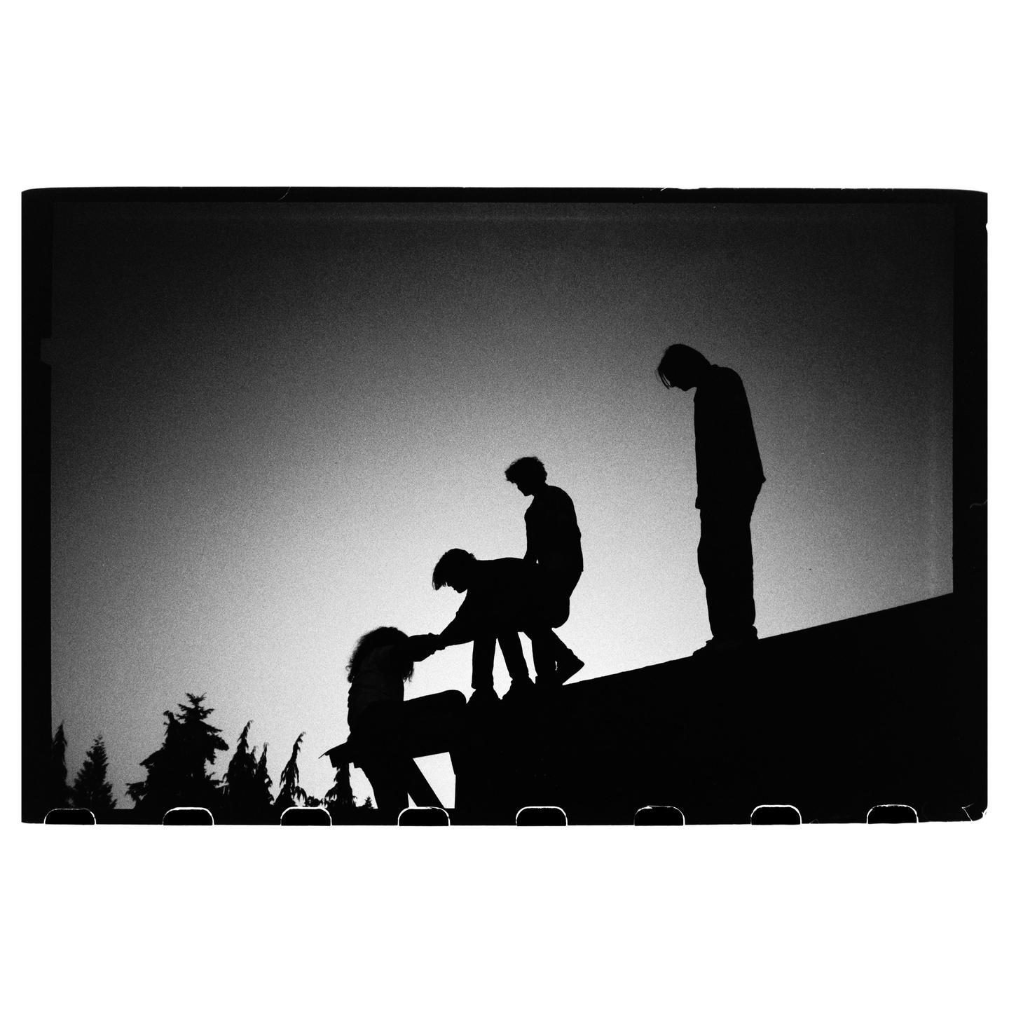 Three men are silhouetted against a bright sky, with one man helping another up a slope.