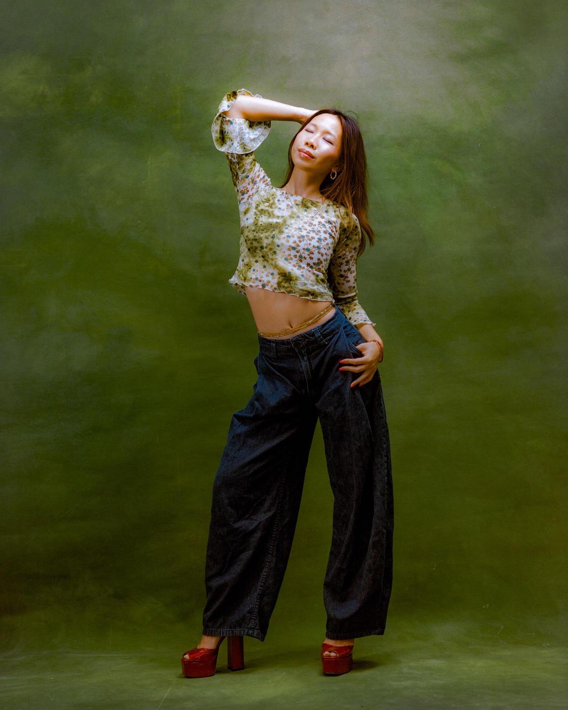 A woman with long hair and closed eyes poses against a green background, wearing a floral crop top, wide-leg jeans, and red platform shoes.