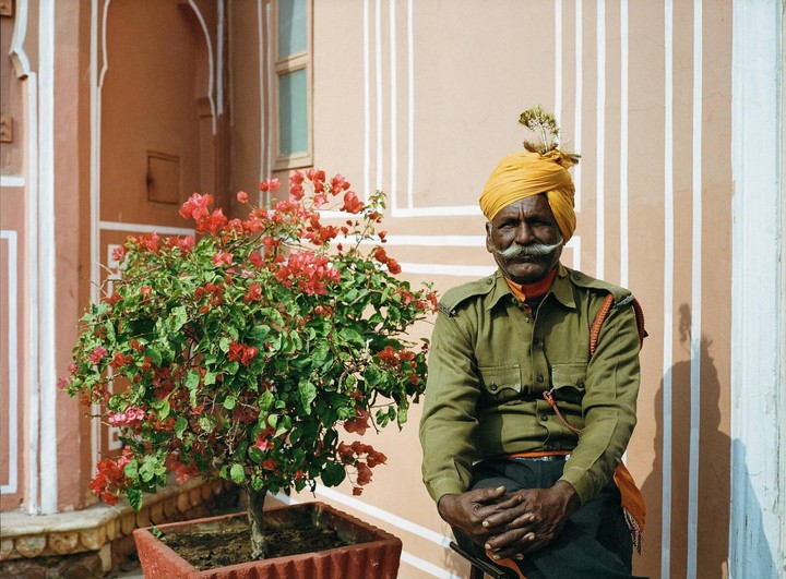 A man in a yellow turban and green uniform sits next to a potted plant with red flowers against a peach-colored wall.
