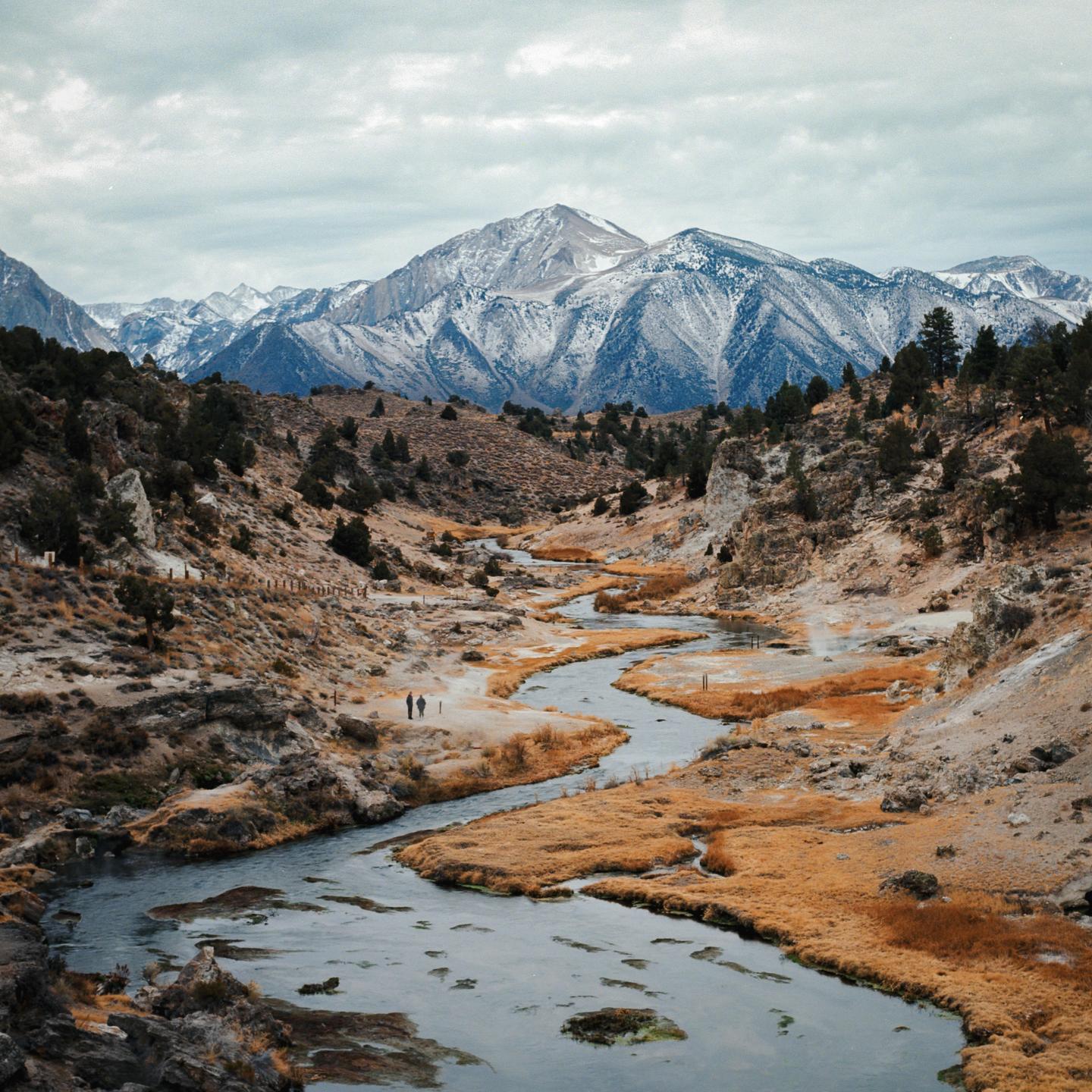 A winding river flows through a valley with two people walking on its bank, leading to snow-capped mountains under a cloudy sky.
