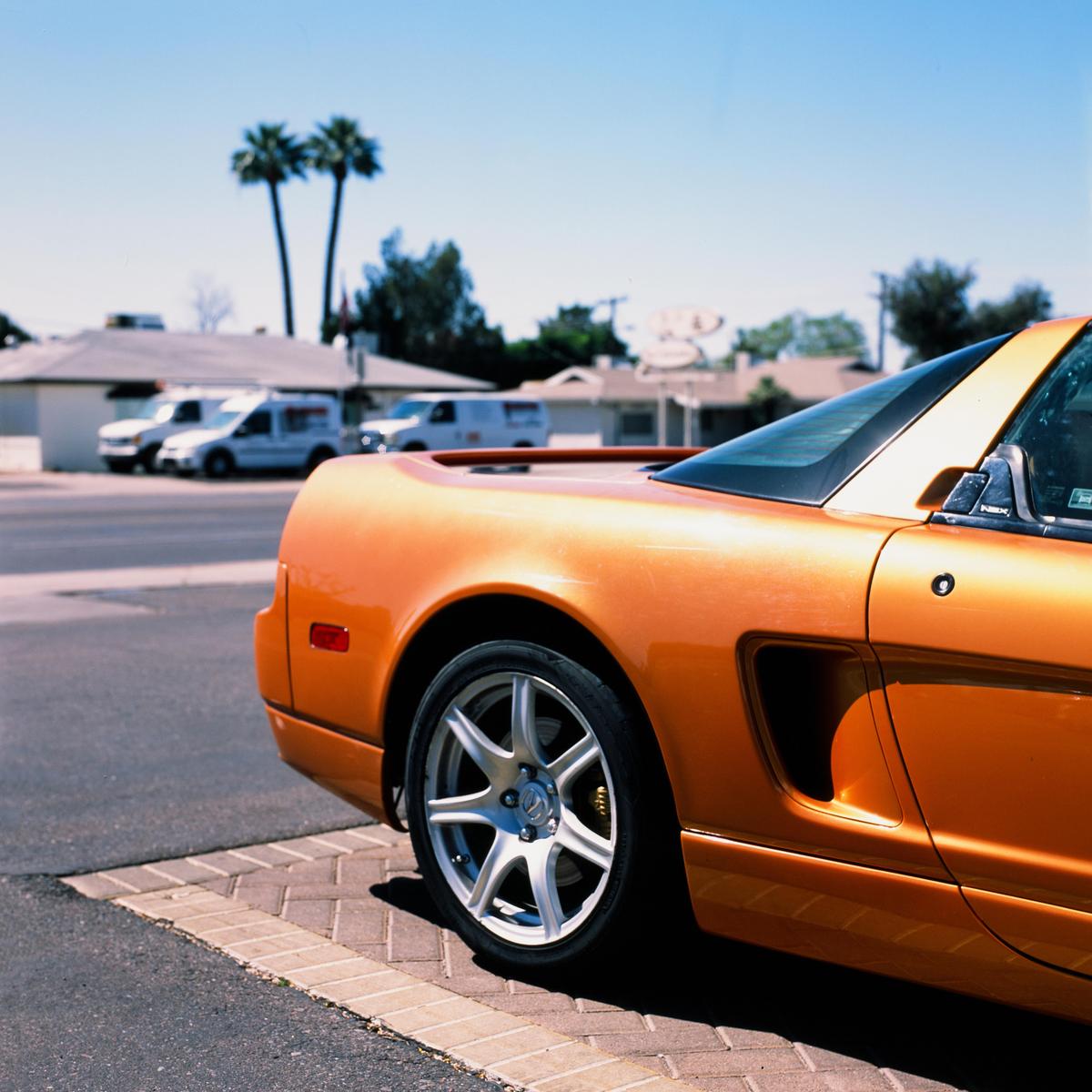 An orange sports car is parked on a paved surface with palm trees and buildings in the background.