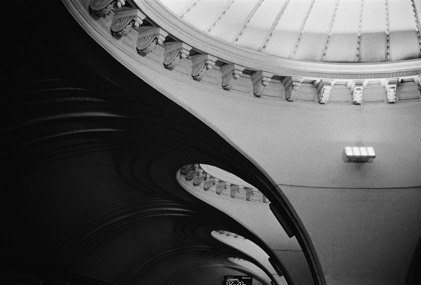 A black and white photo shows the curved architecture of a building interior with decorative elements and a light fixture.