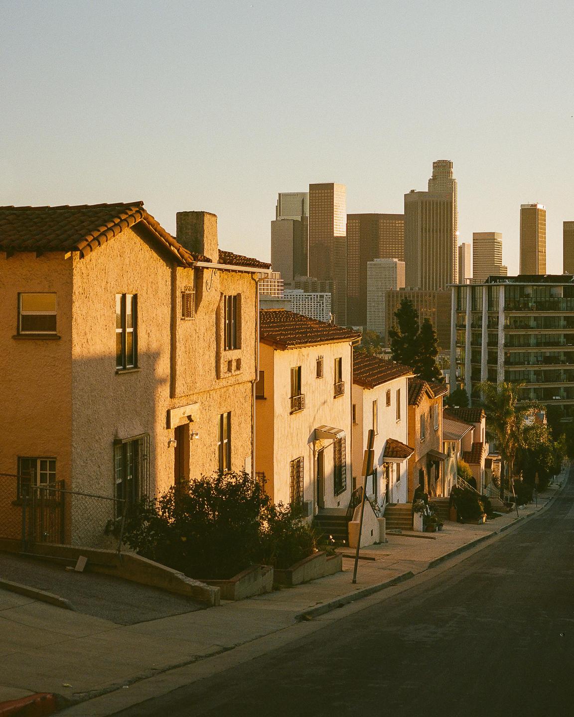A row of houses with terracotta roofs sits on a hill overlooking a city skyline at golden hour.