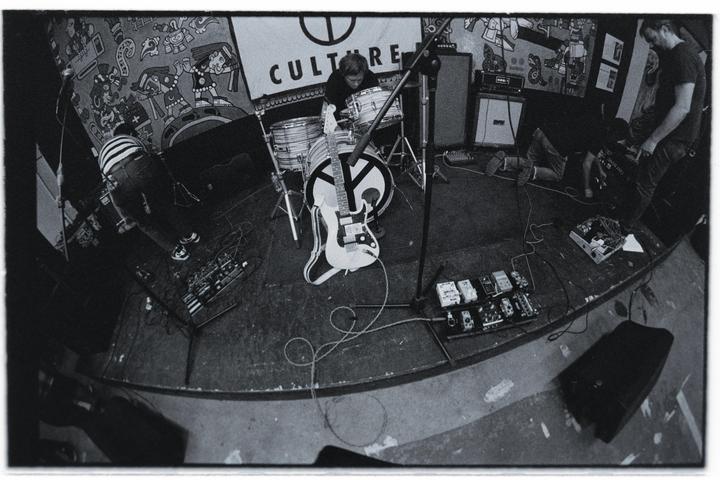 Three men are on a stage with musical instruments and equipment, including a drum kit and an electric guitar.