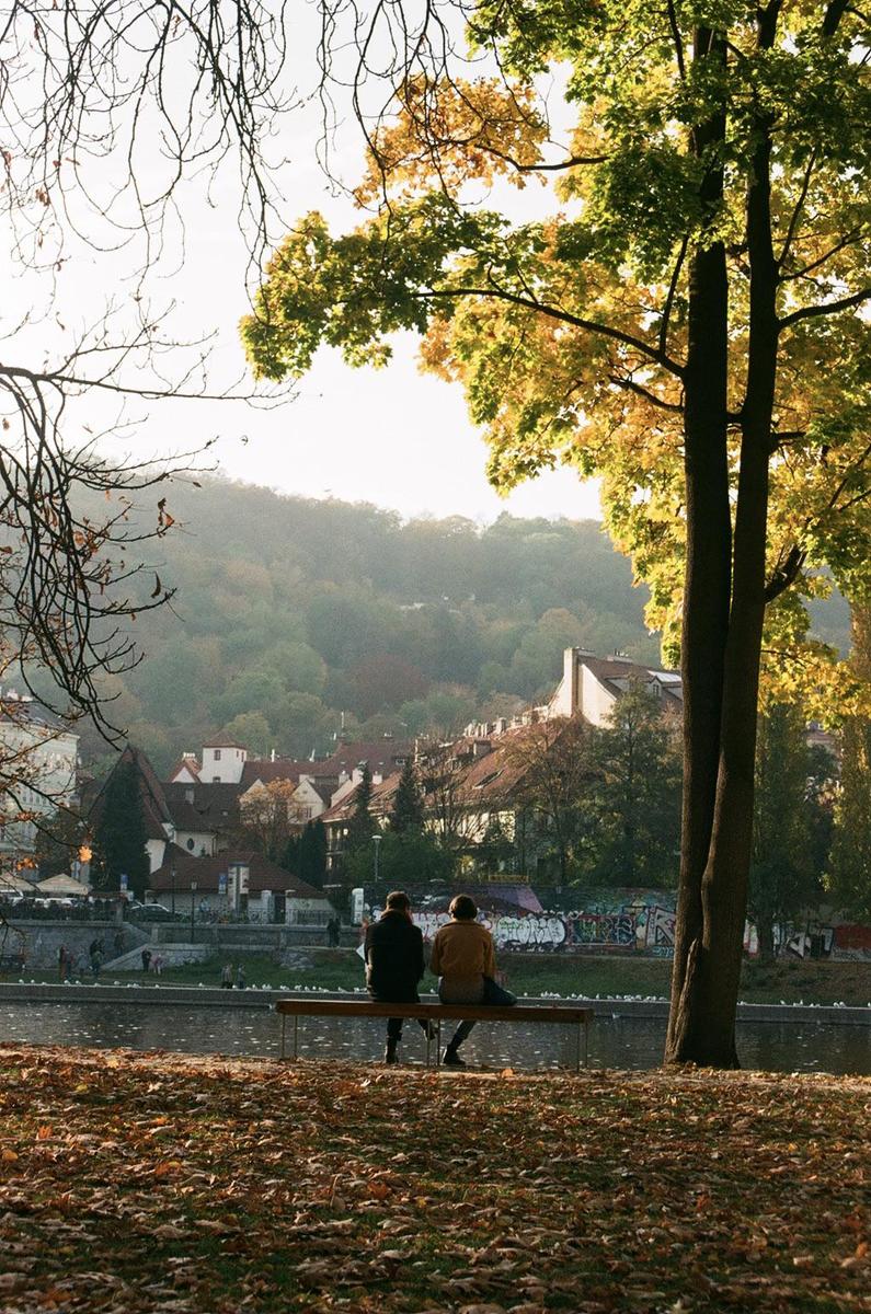 Two people sit on a bench by a river with a town and hills in the background.