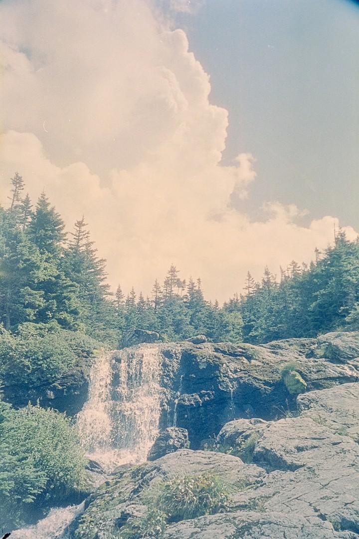 A waterfall cascades over rocks surrounded by evergreen trees under a cloudy sky.