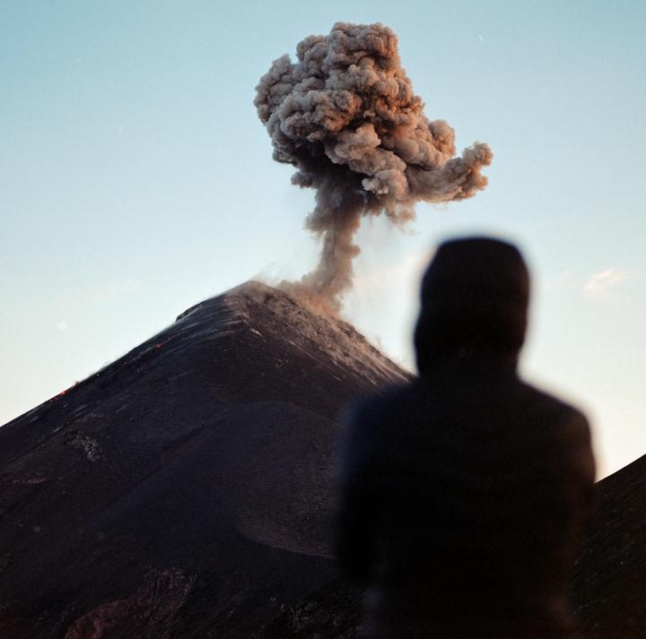 A person stands in the foreground, looking at a volcano erupting smoke and lava.