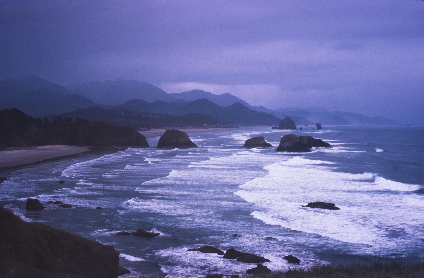 Waves crash on a beach with large rock formations and mountains in the background under a purple sky.