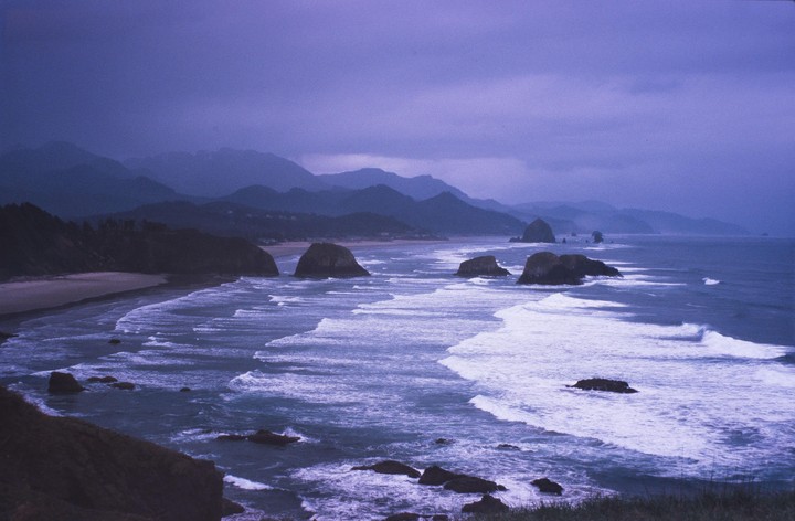 Waves crash on a beach with large rock formations and mountains in the background under a purple sky.