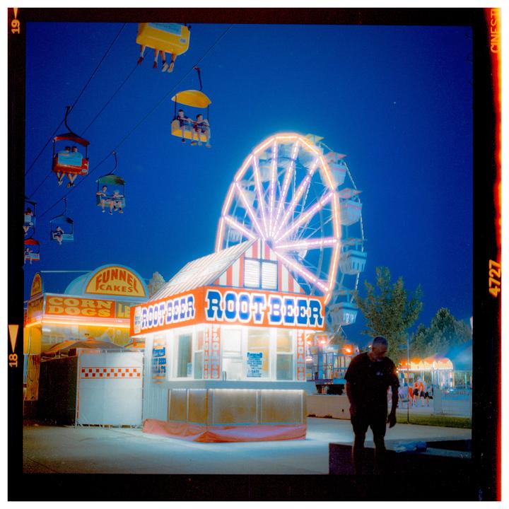 A man stands in front of a root beer stand and a ferris wheel at a fair at night.