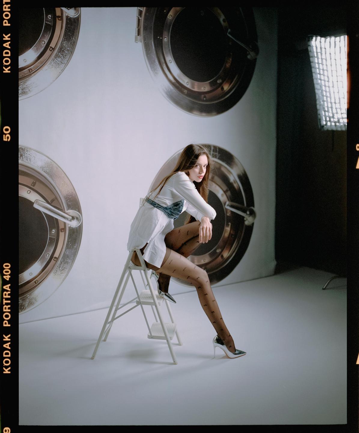 A woman in a white shirt and patterned stockings sits on a stepladder in a studio with circular designs on the wall.