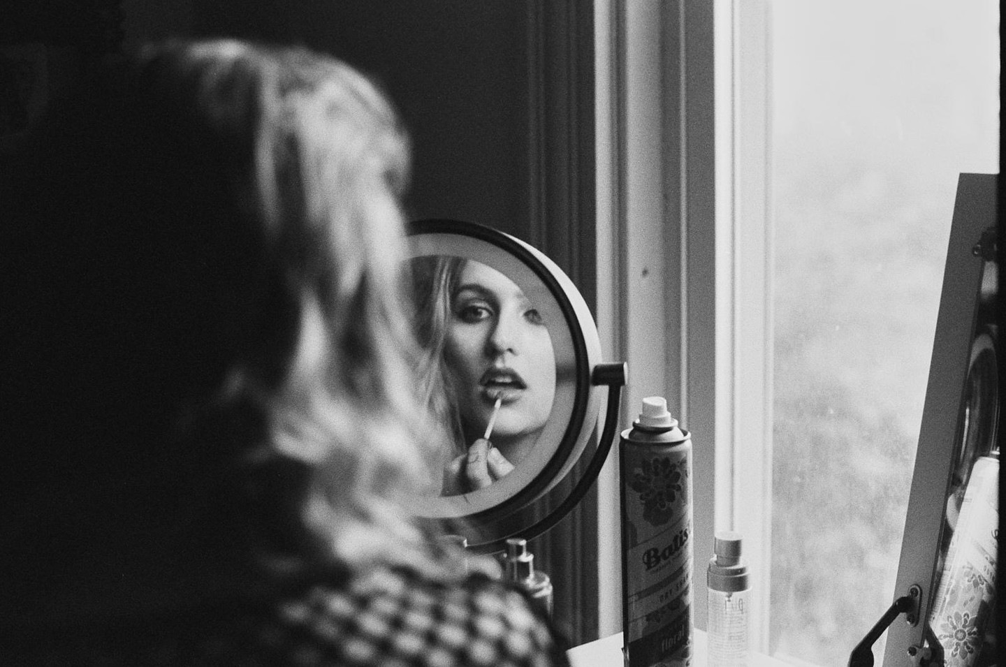 A woman applies lip gloss while looking into a round mirror, with her back to the viewer.